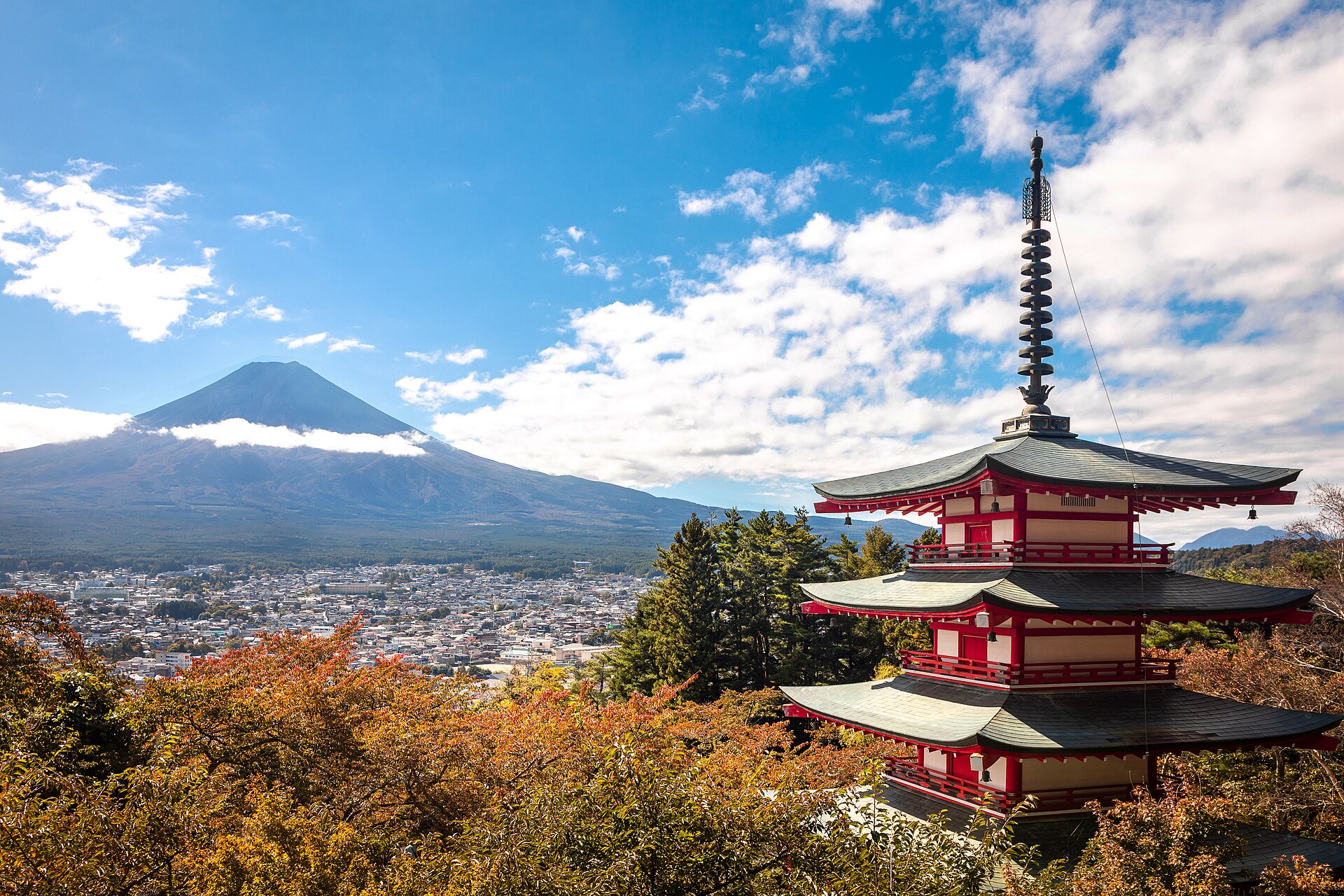 Pięciokondygnacyjna pagoda Chureito na tle miasta Fujiyoshida i Fuji w październiku 2024, na pierwszym planie jesienne liście. Fuji bywa zasłonięta chmurami.