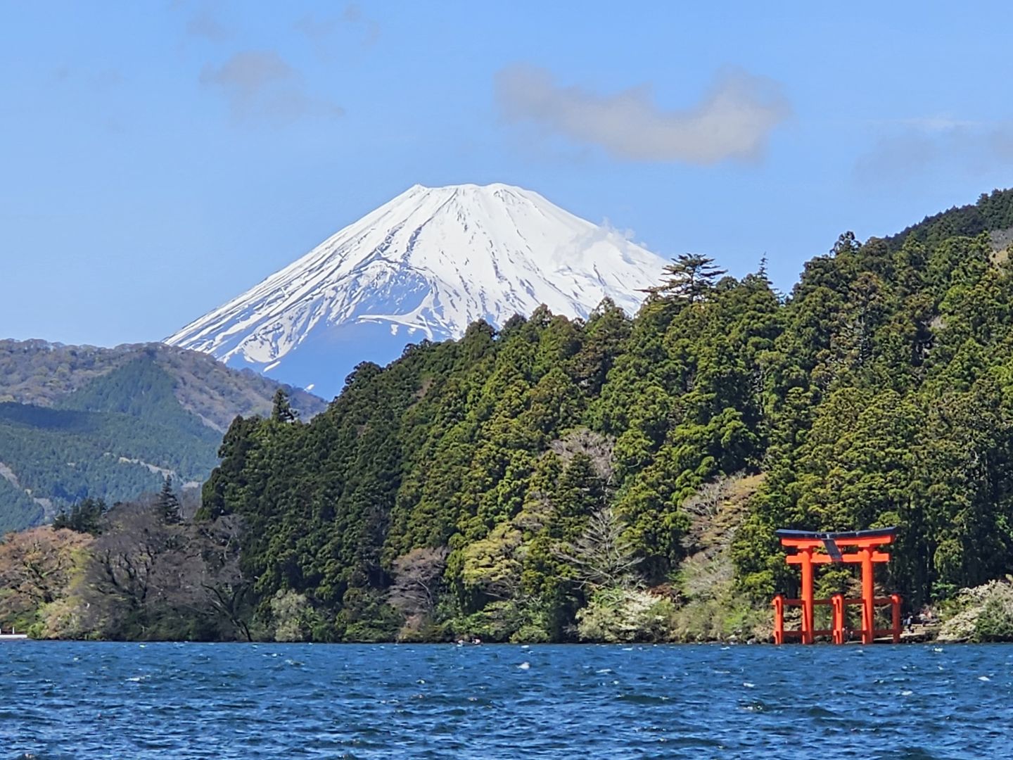 Czerwona Heiwa-no-Torii w wodzie pod zalesionym brzegiem i ośnieżona Fuji w tle, widok od strony Motohakone-ko, kwiecień 2023; Fuji widać tylko przy dobrej widoczności.