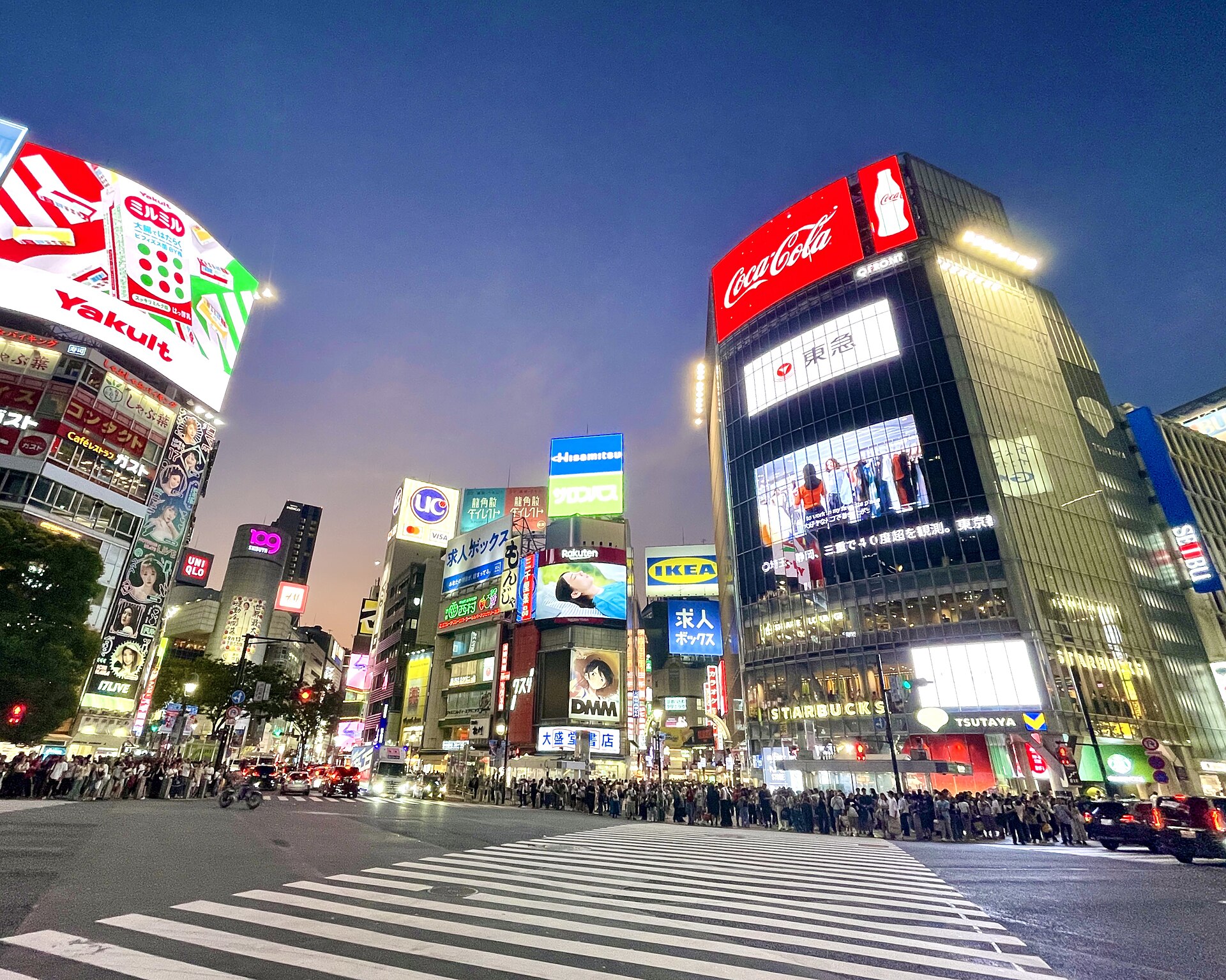 Shibuya Scramble Crossing o zmierzchu z poziomu ulicy: pasy, tłum i neony (m.in. QFRONT). Układ reklam może się zmienić.